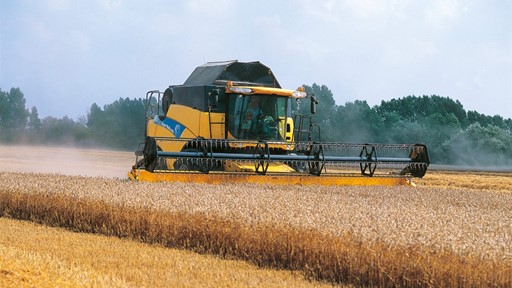 A yellow and black combine harvester working in a golden wheat field under a blue sky with scattered clouds. Dust is being kicked up as the machine moves through the crops, surrounded by green trees in the background.