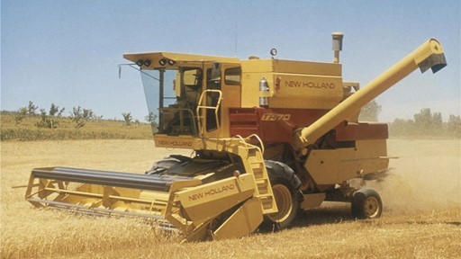A yellow New Holland combine harvester working in a golden wheat field under a clear blue sky. The machine is actively harvesting crops, with dust rising around it, showcasing agricultural machinery in action.
