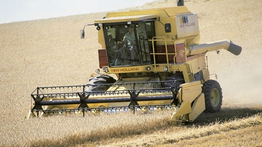 A yellow New Holland combine harvester working in a golden wheat field under a clear blue sky. The machine is cutting through the crops, with a farmer visible inside the cab, showcasing agricultural machinery in action.