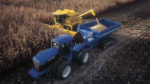 A blue tractor and a yellow combine harvester are working in a cornfield. The tractor is equipped with a large trailer, while the combine is harvesting corn. The landscape features rows of tall corn plants under a cloudy sky, showcasing an agricultural setting during the harvest season.