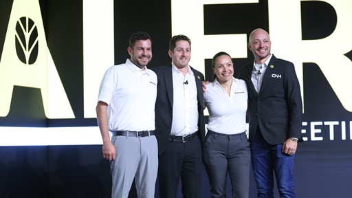 Four professionals pose together on stage at a corporate event with a large sign in the background They are dressed in business attire with two wearing white polo shirts and the others in a suit and a formal shirt The setting suggests a business meeting or conference