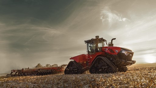A red Case IH tractor with large rubber tracks is working in a harvested field under a dramatic cloudy sky during sunset, pulling farming equipment behind it.