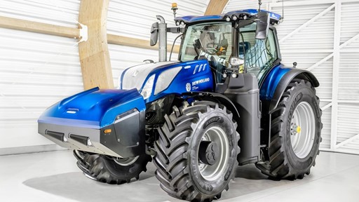 A blue New Holland T7.270 tractor with large black tires is parked inside a bright, modern barn with white walls and wooden beams. The tractor features a spacious enclosed cabin and front weights for balance.