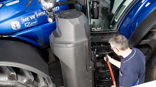 A man in a navy blue shirt is inspecting or repairing the steps of a blue New Holland T7.270 tractor inside a workshop or garage. The tractor's large front tire and side mirror are visible, along with the "Methane Power" label on the tractor's hood.