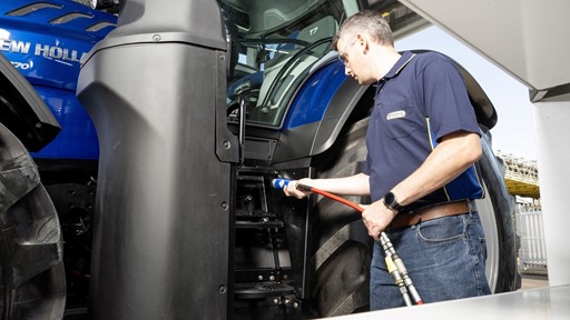 A man wearing a navy blue polo shirt and jeans is inspecting or refueling a large blue New Holland tractor. He is holding a red hose connected to a nozzle near the tractor's steps, standing next to the tractor's large rear tire. The scene appears to be outdoors under a covered area.