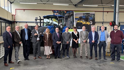 A group of twelve professionally dressed people standing in a row inside an industrial workshop with a large blue tractor behind them. The setting appears to be a formal event or visit, with a mix of men and women posing for a group photo. The background includes industrial equipment, posters, and workshop lighting.