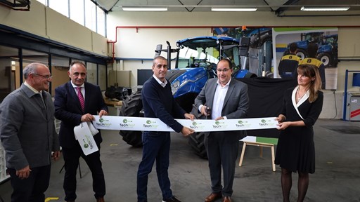 Five people standing in a workshop with a blue tractor in the background, participating in a ribbon-cutting ceremony. One man in a gray suit is cutting a white ribbon with "TechPro" logos, while the others hold the ribbon and smile. The setting appears to be an industrial or agricultural equipment facility.