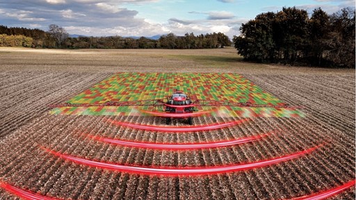 A red agricultural tractor equipped with advanced technology is spraying a field, with a digital overlay showing a colorful grid pattern on the crops and red curved lines representing sensor or radar signals, illustrating precision farming and smart agriculture techniques in a rural landscape under a partly cloudy sky.