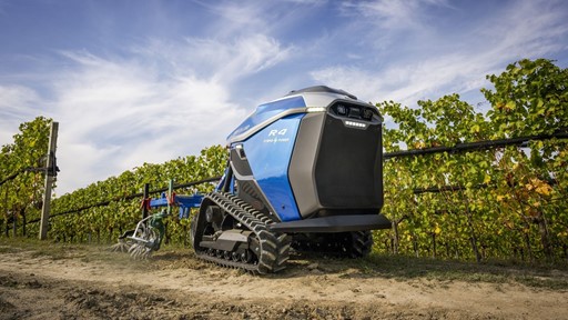 A blue autonomous agricultural robot with tank-like tracks is working on a dirt path next to rows of green grapevines under a partly cloudy sky. The robot is equipped with farming tools and appears to be designed for vineyard maintenance.