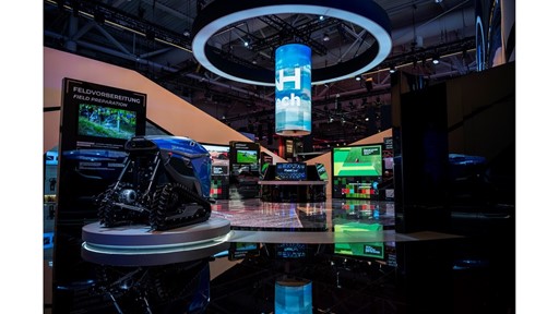 Exhibition booth showcasing advanced agricultural technology with a blue autonomous robotic vehicle on a circular platform, multiple digital screens displaying field preparation and seed planting information, and a large cylindrical hanging digital display with the word "Tech" in a modern indoor event space.
