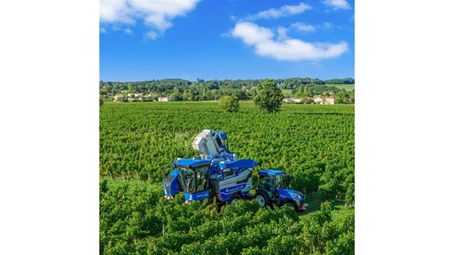 A large blue grape harvester machine and a blue tractor working in a lush green vineyard under a bright blue sky with scattered white clouds, with a backdrop of trees and distant houses.