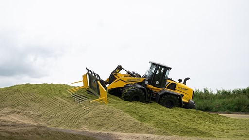 A yellow New Holland W170D wheel loader is pushing and leveling a large pile of green silage on a farm, with a cloudy sky in the background.