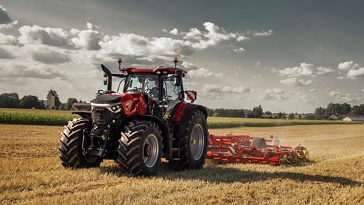 A red Case IH tractor with large tires is working in a golden wheat field under a partly cloudy sky, pulling a red agricultural implement for tilling the soil, with green crops and trees visible in the background.