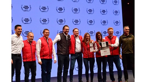 A group of nine people standing on stage in front of a large screen displaying the "Farm Machine 2026" logo repeatedly. Most individuals are wearing red vests over white shirts, while two are in different colored jackets. Two women in the center are holding awards, one holding a plaque and the other a certificate. The group is smiling and posing for a photo, suggesting a celebratory or award event.