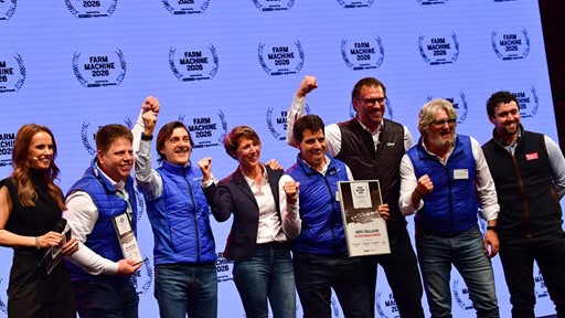 A group of eight people standing on stage celebrating an award at the Farm Machine 2026 event. Most of them are wearing blue vests and smiling, with some raising their fists in excitement. One person is holding a framed certificate that reads "New Holland Methane Power." The background features a repeated "Farm Machine 2026" logo.