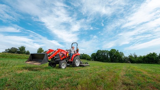 A man driving a red tractor with a front loader attachment across a green grassy field under a partly cloudy blue sky, with trees in the background.