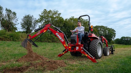 A man operating a red CASE IH tractor equipped with a backhoe attachment, digging into the grassy ground in a rural outdoor setting with trees and a cloudy sky in the background.