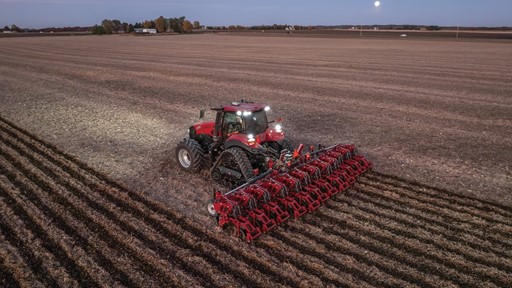 A red tractor with large tracks is plowing a vast, open field at dusk, creating evenly spaced furrows in the soil under a clear sky with the moon visible in the background.
