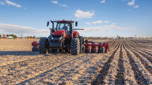 A red tractor with large tires is plowing a vast, dry agricultural field under a blue sky with scattered clouds. The tractor is positioned in the center of the image, moving towards the camera, creating evenly spaced furrows in the soil. Trees and farm buildings are visible in the distant background.