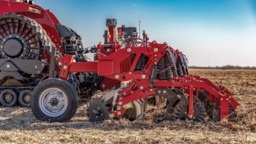 Close-up view of a red modern agricultural tractor attachment with multiple disc blades and wheels working on a dry field under a clear blue sky, preparing the soil for planting.