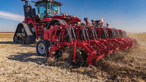 A large red agricultural tractor with rubber tracks is pulling a multi-row planter attachment across a dry field under a clear blue sky, preparing the soil for planting.