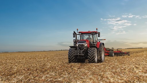 A red tractor with large tires is working in a vast harvested cornfield under a clear blue sky with scattered clouds during sunset.