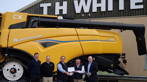 Five men standing in front of a large yellow New Holland CX combine harvester outside a building with a sign that reads "TH WHITE." Two of the men in the center are shaking hands and holding a certificate, while the others stand beside them smiling.