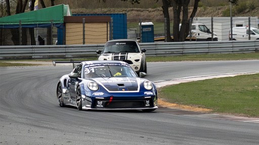 Two race cars navigating a curved section of a racetrack, with a blue and white Porsche leading and a white Mini Cooper with black racing stripes following closely behind, surrounded by safety barriers and grassy areas.
