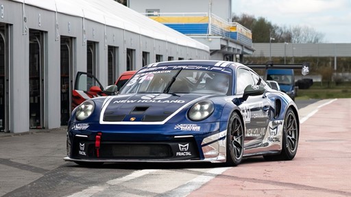 A blue and white Porsche race car with the number 31 and "New Holland" branding is parked on a race track pit lane next to a row of garages. The car features racing decals, a large rear wing, and a red tow hook on the front bumper. The background shows a partly cloudy sky and some greenery.