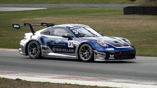 A blue and silver Porsche race car with number 31 on the door is driving on a race track. The car features sponsor logos including Orvanthe Performance, New Holland, XPEL, and Fractal, and has a large rear wing for aerodynamics. The background shows grassy areas and tire barriers along the track.