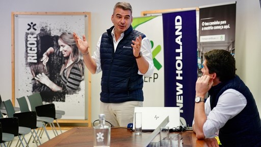 Two men in a meeting room, one standing and speaking with expressive hand gestures while the other sits at a table listening attentively. Both are wearing navy blue vests over white shirts. The table has laptops, glasses of water, and a water bottle. In the background, there are banners with the logos "NEW HOLLAND" and "RIGOR," and a poster featuring a woman working with her hands. The setting appears to be a professional or corporate environment.