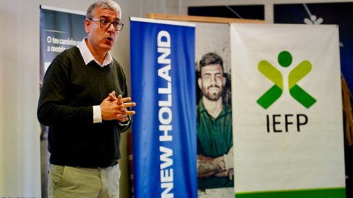 A man with gray hair and glasses, wearing a black sweater over a white collared shirt and beige pants, is speaking or presenting indoors. Behind him are three vertical banners, one blue with the text "NEW HOLLAND," another with a photo of a man with crossed arms, and a white banner with a green and yellow logo and the text "IEFP.