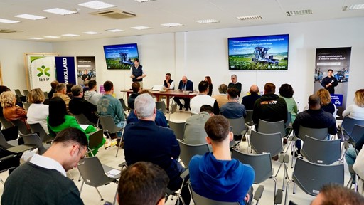 A group of people attending a presentation or seminar in a conference room. A speaker stands at the front near two large screens displaying slides with agricultural machinery images and text. Several panelists are seated at a table beside the speaker. Attendees are seated in rows facing the front, some taking notes or using their phones. Banners for IEFP and New Holland are visible in the background. The setting appears professional and focused on agricultural or farming topics.