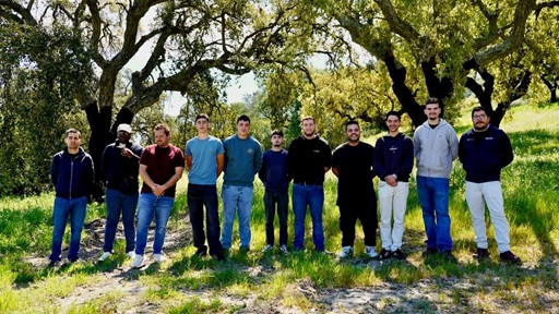 A group of ten young men standing outdoors in a sunny, grassy area with large trees in the background, all facing the camera and posing for a group photo.