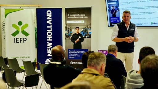 A man in a white shirt and black vest is giving a presentation to an audience seated in a room. Behind him are banners for IEFP and New Holland, and a screen displaying information in Portuguese about a training course and collaboration between IEFP and New Holland. The audience is attentively listening, and the setting appears to be a professional or educational event focused on agricultural machinery training.