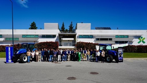 A large group of people standing in front of a modern white building with green bushes and trees in the background. Two blue New Holland tractors are positioned on either side of the group, with New Holland banners next to each tractor. The sky is clear and blue, indicating a sunny day.