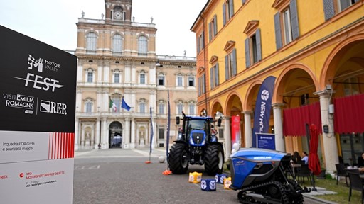 Outdoor exhibition of New Holland agricultural machinery including a blue tractor and a tracked vehicle displayed on a cobblestone street in front of historic buildings with arches and a clock tower in the background, during the Motor Valley Fest in Emilia Romagna, Italy.