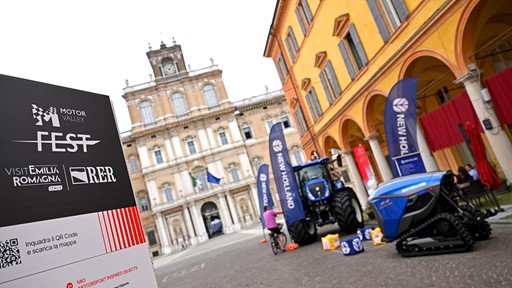 Outdoor event setup in a historic Italian square featuring a Motor Valley Fest sign with logos of Visit Emilia Romagna and RER, alongside New Holland branded agricultural machinery including a blue tractor and a blue tracked vehicle, with a person on a bicycle nearby and classical architecture in the background.