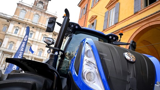Close-up view of a blue New Holland tractor displayed outdoors in front of historic European-style buildings with arched windows and shutters, under a partly cloudy sky.