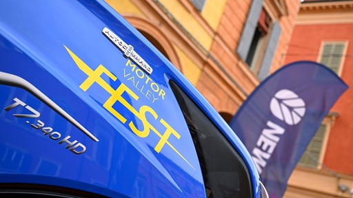 Close-up of a blue vehicle with yellow text "Motor Valley Fest" and model designation "T7.340 HD" on its side, set against a background of colorful buildings and a blurred flag with the word "New" visible.