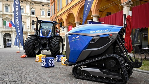 A blue New Holland R4 Hybrid Power autonomous agricultural vehicle with tracks is displayed on a cobblestone street next to a traditional blue New Holland tractor with large tires. The scene is set in an urban area with historic buildings and arches in the background, accompanied by New Holland branded flags and colorful cube seats on the ground.