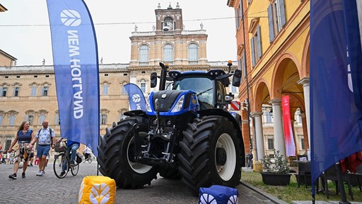 A large blue New Holland tractor is displayed on a cobblestone street in front of historic buildings with arched windows and a clock tower. Blue New Holland flags and branded cube cushions are placed around the tractor. People walk and ride bicycles nearby, enjoying the outdoor event.