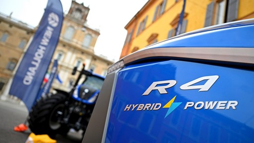 Close-up view of the front side of a blue R4 Hybrid Power tractor with a blurred background showing another tractor, flags, and historic buildings in an outdoor urban setting.