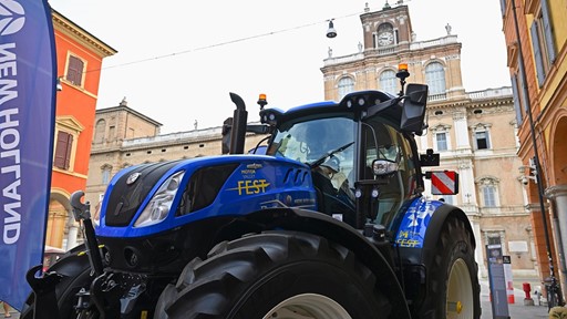 Blue New Holland tractor with "Motor Valley Fest" branding displayed in an urban setting with historic buildings in the background, under a clear sky.
