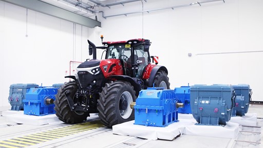 A red Case IH tractor is positioned indoors on a testing platform surrounded by several large blue industrial gearboxes and machinery components, all set in a clean, well-lit white room with visible ceiling pipes and safety markings on the floor.