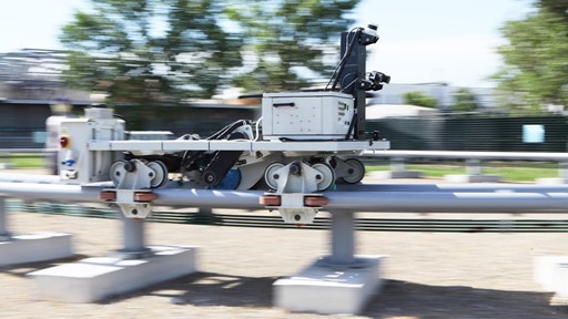 A mobile robot equipped with cameras and sensors is moving along a metal rail outdoors, with blurred trees and buildings in the background indicating motion.