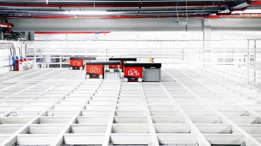 A high-tech automated warehouse with red robotic carts numbered 018, 020, and 026 moving along white grid-like storage shelves under a ceiling with red pipes and industrial lighting.