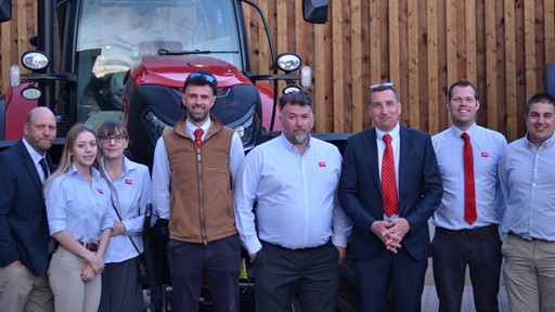 A group of eight professionally dressed people standing in front of a large red tractor and a wooden wall, posing for a group photo. Most individuals are wearing light blue shirts, some with red ties, and one person is wearing a brown vest.