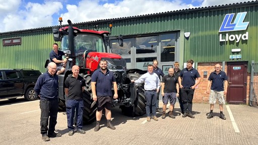 A group of nine men standing and posing in front of a large red tractor outside a green industrial building with the sign "Lloyd" on it. One man is sitting on the tractor, while the others stand on the ground, smiling and dressed in casual work attire. The scene is set on a sunny day with a blue sky and some clouds.