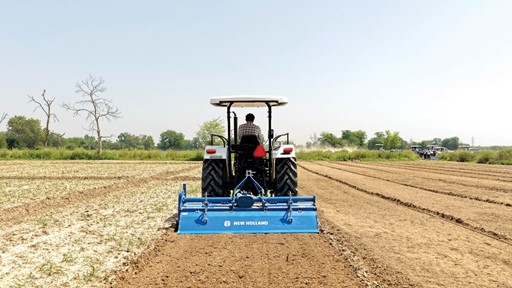 A farmer driving a white New Holland tractor with a blue tiller attachment, plowing a large, dry agricultural field under a clear sky, with green trees and other tractors visible in the background.